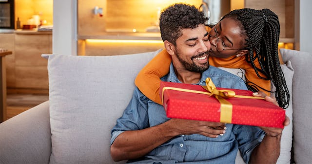 A woman handing her husband a meaningful holiday gift wrapped in red decorative paper while he's sit...