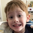 An autistic boy with brown hair and a blue shirt smiling at the camera in his room