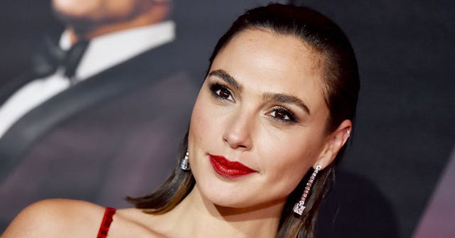 Gal Gadot posing during a red carpet event while sporting a red dress, red lipstick and silver earri...