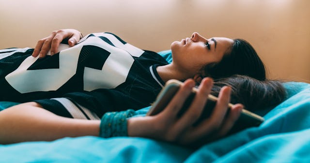 Black-haired girl wearing a black shirt while lying on a blue bed and holding her mobile phone