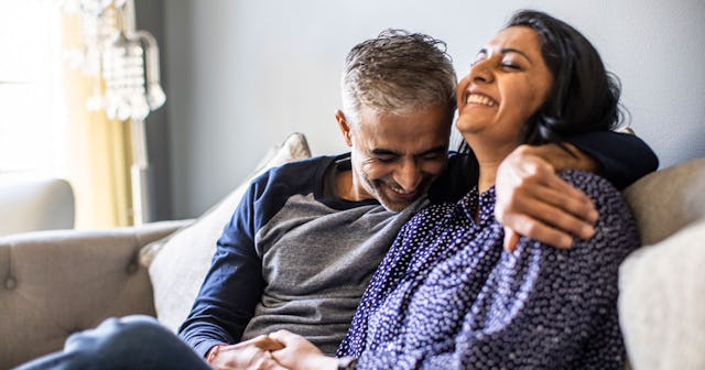 Partners in a middle-aged marriage, smiling while sitting hugged on the couch inside their home.