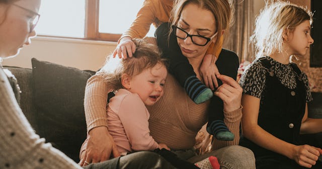 A working mother sitting on a couch with four kids with one crying and one climbing over her back