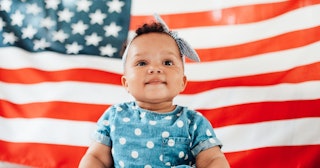 A toddler sits in front of an American flag.