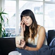 A tired-looking woman sits on the sofa, holding a cup of coffee and rubbing her eye during the pande...