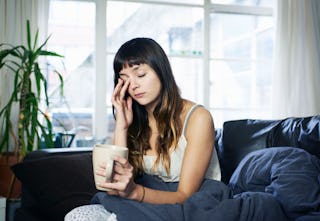 A tired-looking woman sits on the sofa, holding a cup of coffee and rubbing her eye during the pande...