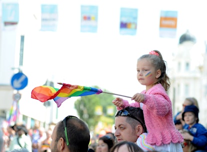 A child holding a pride flag with a rainbow painted on her face, sitting on her fathers shoulders at...