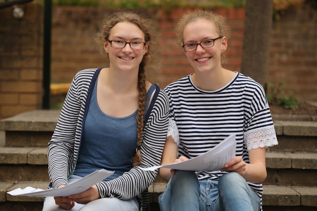 Two girl-friends sitting on stairs and holding papers are smiling at the camera.