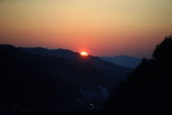 A view of a landscape during the sunset with the sun visible behind multiple mountains