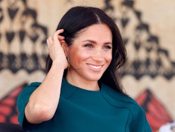 Meghan Markle in a teal dress smiling as she's moving her hair away from her face with black nail po...