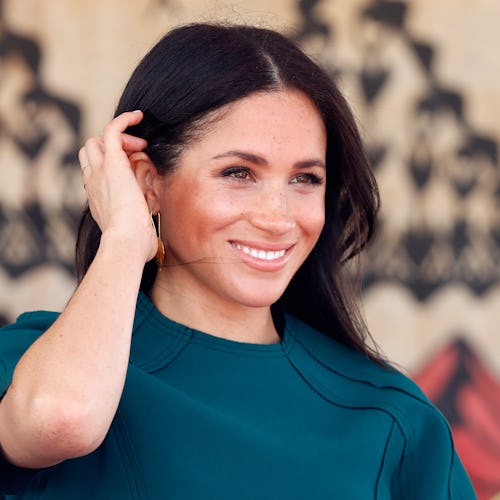 Meghan Markle in a teal dress smiling as she's moving her hair away from her face with black nail po...