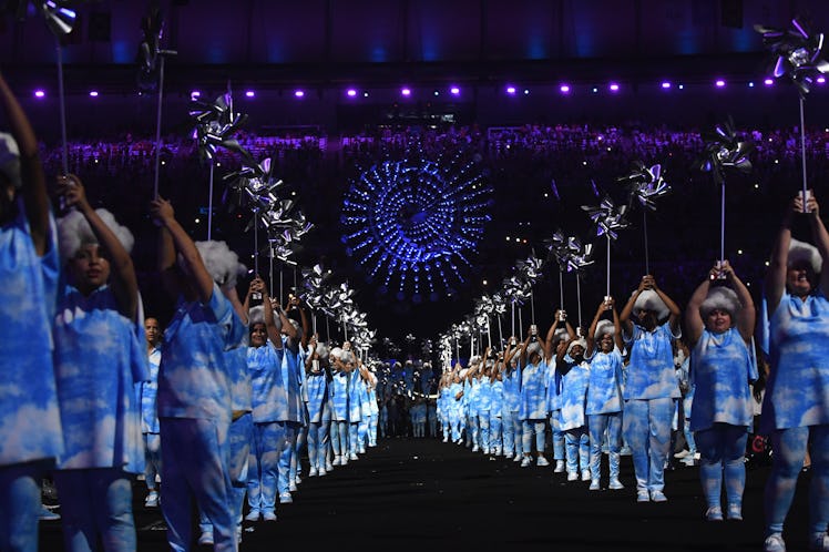 Performers wearing outfits decorated with blue and white clouds hold pinwheels in front of a blue sp...