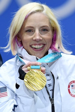 Kikkan Randall in a white U.S. Olympic Team jacket, smiling while holding up a gold medal