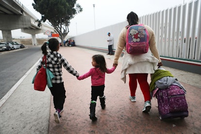 An immigrating family of three where mom is holding hands with her daughters and pulling a suitcase