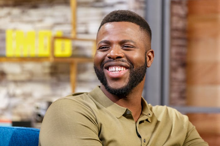 Winston Duke in a beige polo shirt, laughing on a TV show