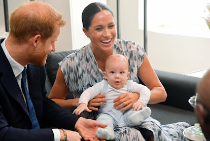 Meghan Markle in a dress, holding Archie in her lap and laughing, as Prince Harry sits next to them ...