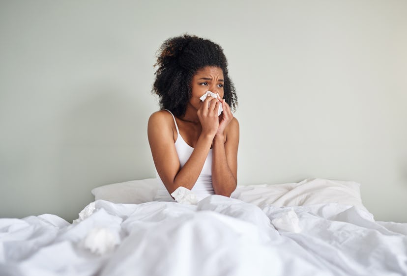 A woman sneezes in bed. Having the flu and your period at the same time is unpleasant, and periods m...