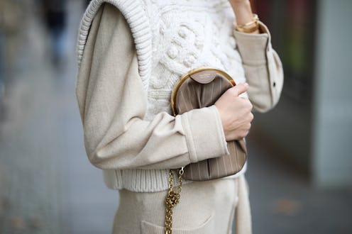 A woman wearing a white knitted sweater over a beige shirt holds a brown handbag to her