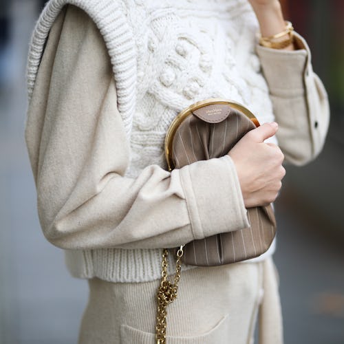A woman wearing a white knitted sweater over a beige shirt holds a brown handbag to her