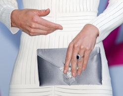 A woman in a white dress, holding a silver clutch while wearing a non-diamond engagement ring. 