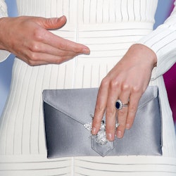 A woman in a white dress, holding a silver clutch while wearing a non-diamond engagement ring. 