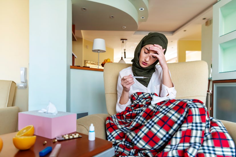 A person wrapped in a plaid blanket sits looking at a thermometer with tissues and oranges on the ta...