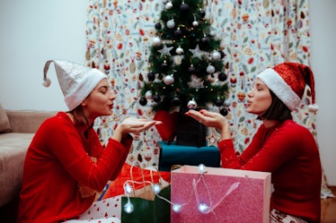 Two women wearing red sweaters and Santa hats smile and get ready to blow fake snow over their prese...