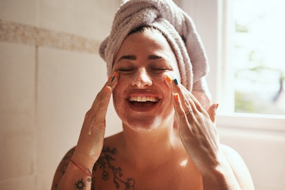 A girl with tattoos and a towel on her head laughs while applying an at-home facial.