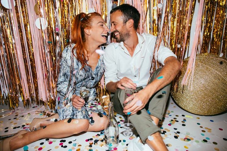 A couple sits on a floor covered with confetti on New Year's Eve as they toast their champagne flute...