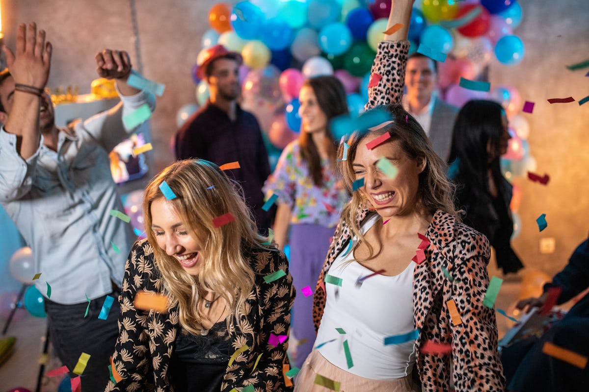 Two blonde girls smile and party while confetti falls from ceiling on New Year's Eve.