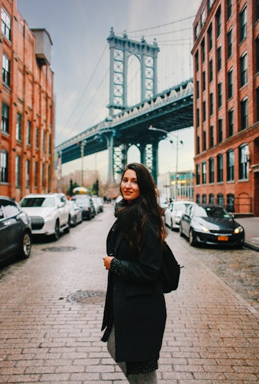 A chic woman poses with the view of the Brooklyn Bridge and warehouses in Dumbo, Brooklyn.