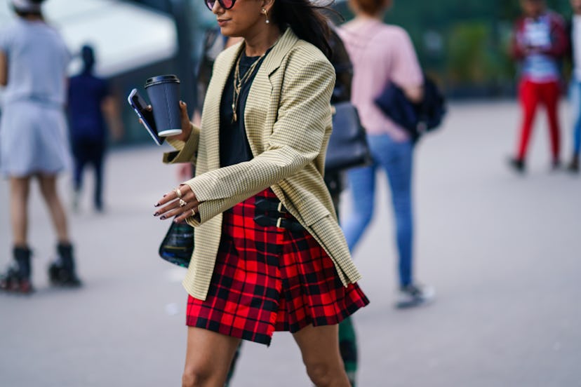 A woman in a plaid skirt, beige blazer and a black shirt holding a coffee cup 