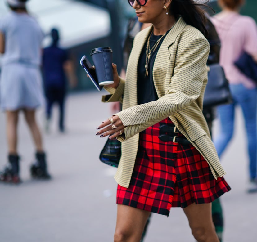 A woman in a plaid skirt, beige blazer and a black shirt holding a coffee cup