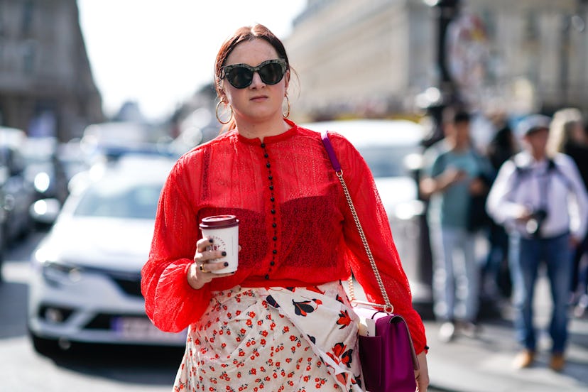A woman in a sheer red top, and sunglasses walking down the street with a coffee cup in her hand 