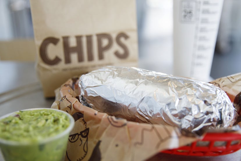 A Chipotle burrito sits in foil next to a side of guacamole and a bag of chips in the background. If...
