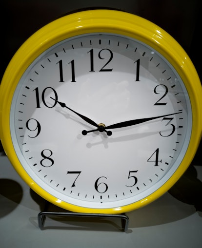 A yellow analogue clock sits on a stand. Give yourself less time to complete a task to help accompli...