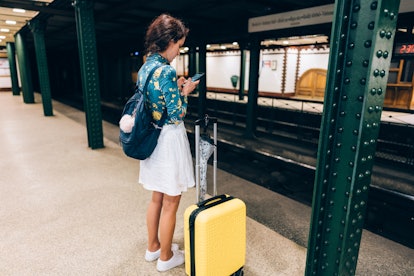A woman stands in a subway station in a city with a yellow suitcase by her side.