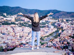 A blonde woman stands on a rock overlooking a city with her arms wide open.