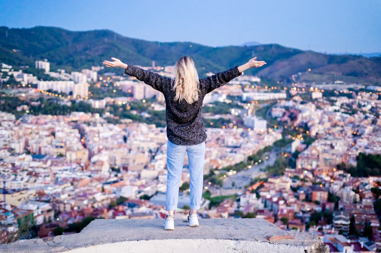 A blonde woman stands on a rock overlooking a city with her arms wide open.