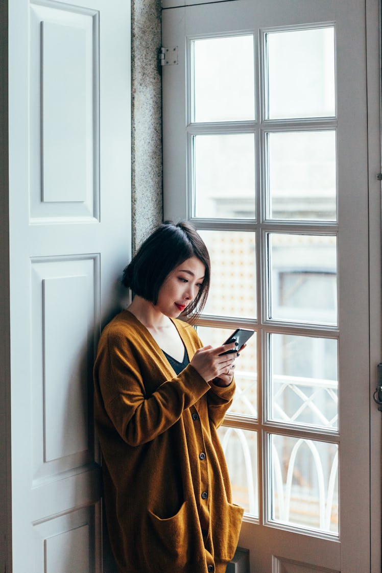 A woman stands in a sunny window while looking at the best travel apps on her phone.