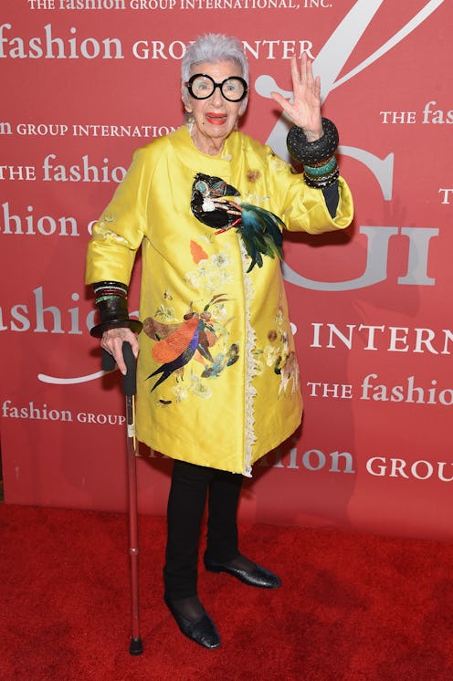 Iris Apfel wears a satin yellow coat with a lace trim adorned with tropical birds of varying texture...