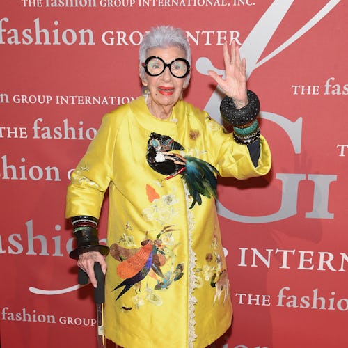 Iris Apfel wears a satin yellow coat with a lace trim adorned with tropical birds of varying texture...