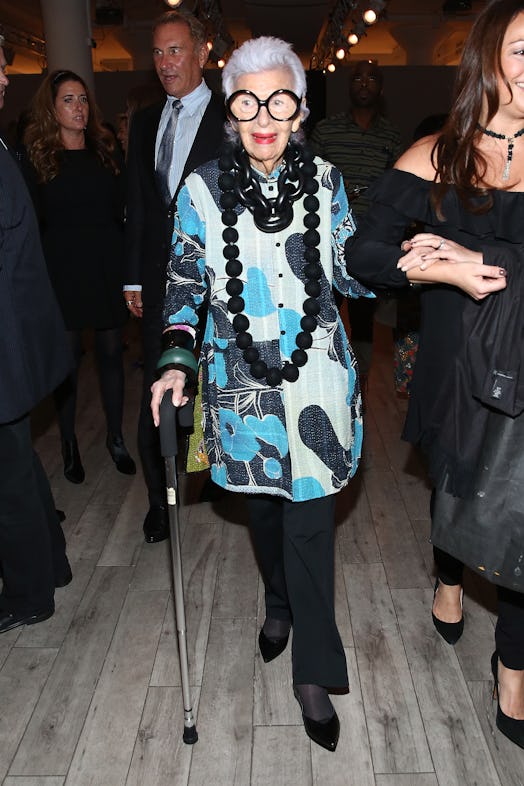 Iris Apfel wearing button up blue printed dress while walking down carrying her crutch