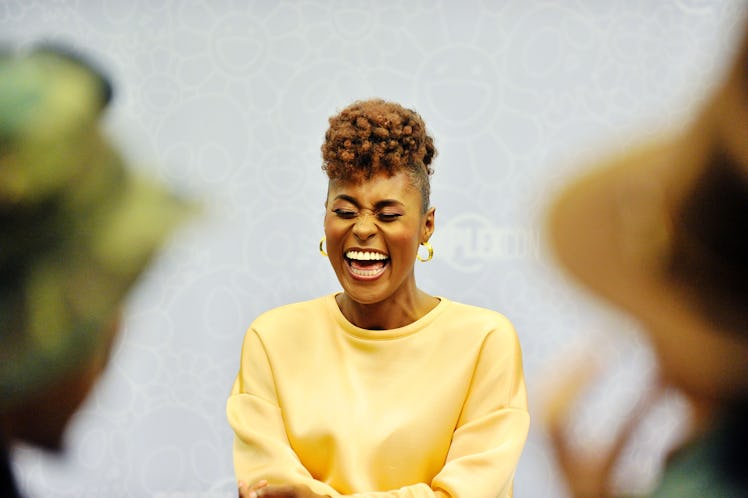 Issa Rae laughing with closed eyes, wearing a yellow sweater and golden hoop earrings