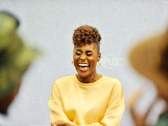 Issa Rae laughing with closed eyes, wearing a yellow sweater and golden hoop earrings