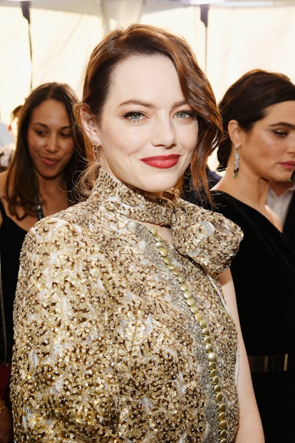 Emma Stone with a gold cat eye, a tousled updo posing in a gold embellished dress 