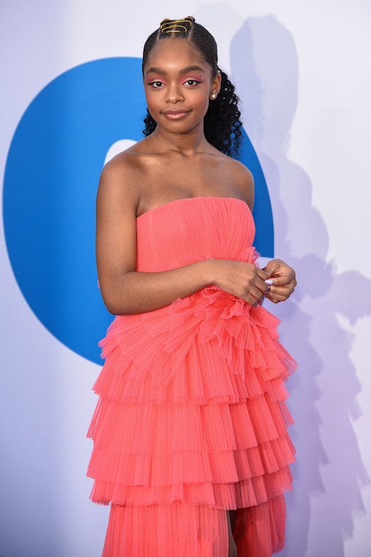 Marsai with a low pony with gilded center braids, wearing a peach-colored frilled dress on the red c...