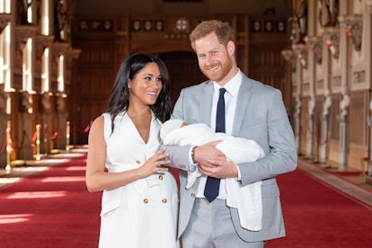 Harry in a formal suit and Meghan in a white dress posing with baby Sussex