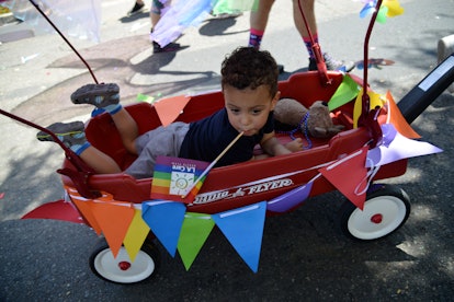 A toddler in a red wagon that's decorated with rainbow colors being pulled at a pride event
