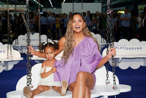 Chrissy Teigen wearing a purple dress next to her daughter Luna with pigtails, in a white dress, on ...