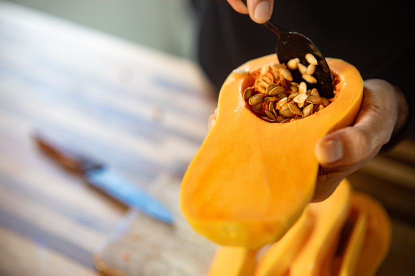 A close up image of a person's hands, holding half of a winter squash while using a spoon to scoop o...